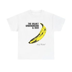 May include: White t-shirt featuring a yellow banana graphic with black accents. The text "THE VELVET UNDERGROUND & NICO" is above the banana, and "Andy Warhol" is below. The shirt is a classic crew neck.
