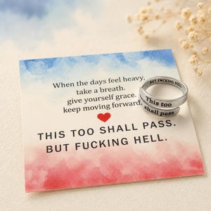 May include: A silver-toned metal ring with the engraved text "This too shall pass" and "BUT F*CKING HELL" resting on a square card. The card features a blue and red watercolor background with the printed text "When the days feel heavy, take a breath, give yourself grace, keep moving forward" and a small red heart.