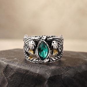 May include: A silver-toned metal ring featuring an oxidized finish and a central marquise-cut emerald green gemstone. The band is designed with intricate relief detailing that resembles stylized bird wings flanking the central stone.