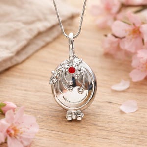 May include: A silver-colored pendant necklace with a red gemstone in the center. The oval-shaped pendant has decorative floral and swirl designs. The necklace is displayed on a wooden surface with pink flowers in the background.