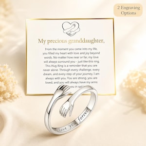 May include: A silver-toned metal ring featuring a design of two hands wrapping around the band in a hug. The band is engraved with the text "Love you forever". The ring is displayed in front of a white card with a heart icon and a printed message.