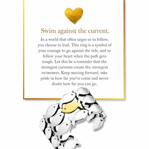 May include: A silver-toned ring featuring a repeating pattern of small fish, with one central fish finished in gold-tone. The ring is displayed in front of a white card with a gold heart icon and the text: Swim against the current. In a world that often urges us to follow, you choose to lead.
