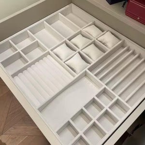 Puede incluir: Un organizador de joyas blanco con múltiples compartimentos. El organizador incluye secciones para anillos, relojes y otras joyas. Los compartimentos están forrados con un material suave de color claro, y algunas secciones tienen pequeñas almohadas cuadradas.