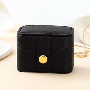 May include: A small, black jewellery box with a textured surface. The box is rectangular with a hinged lid and a gold-coloured button clasp. The box is closed and appears to be empty. The background is a light colour.