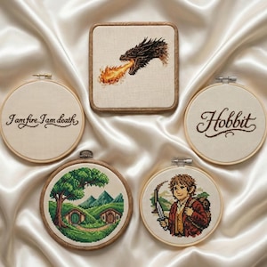 Può includere: Una collezione di opere d'arte a punto croce con immagini de Il Signore degli Anelli e Lo Hobbit. I pezzi includono un drago che sputa fuoco, la parola "Hobbit" e citazioni. I pezzi sono esposti su una superficie di raso bianco.