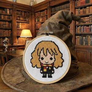 Puede incluir: Un bordado de punto de cruz de un personaje de dibujos animados con cabello rubio, una bufanda roja y dorada y una túnica negra, colocado en un aro de madera. El bordado se coloca frente a un sombrero de mago marrón y desgastado. El fondo es una biblioteca.