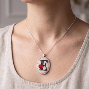 Puede incluir: Collar plateado con un colgante ovalado. El colgante presenta un diseño de punto de cruz con la letra "E" en negro y un corazón rojo. El collar lo lleva una persona.