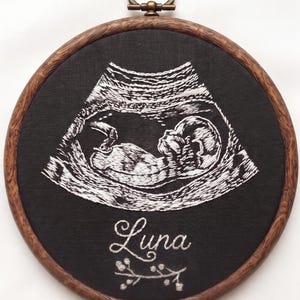 May include: A circular embroidery hoop with a faux wood frame containing black fabric. White thread depicts a fetal ultrasound image. Below the ultrasound, the name Luna is embroidered in white script, followed by a small embroidered sprig design.