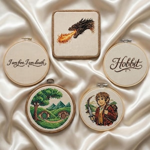 May include: A collection of cross-stitch art pieces featuring scenes and quotes from the fantasy world. Includes a dragon breathing fire, the word "Hobbit", and a character holding a knife. The pieces are in wooden hoops and a square frame.