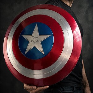 Op de afbeelding: Een rood, wit en blauw schild met een centrale blauwe cirkel en een witte ster. Het schild heeft een zilverkleurige metalen rand. Het wordt vastgehouden door een persoon die een zwart shirt en een blauwe jeans draagt. Een replica van Captain America's schild.