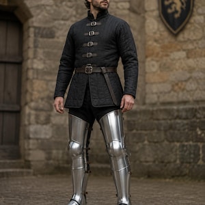 May include: A medieval-style outfit featuring a black quilted jacket with leather straps and a brown belt. The outfit includes shiny silver leg armor. The background shows a stone building and a coat of arms.