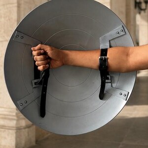 Op de afbeelding: Een grijze, cirkelvormige schild met radiale groeven en metalen beugels. Zwarte leren riemen bevestigen het schild aan de arm. Het schild wordt door een hand vastgehouden, wat duidt op een rekwisiet of kostuumonderdeel.