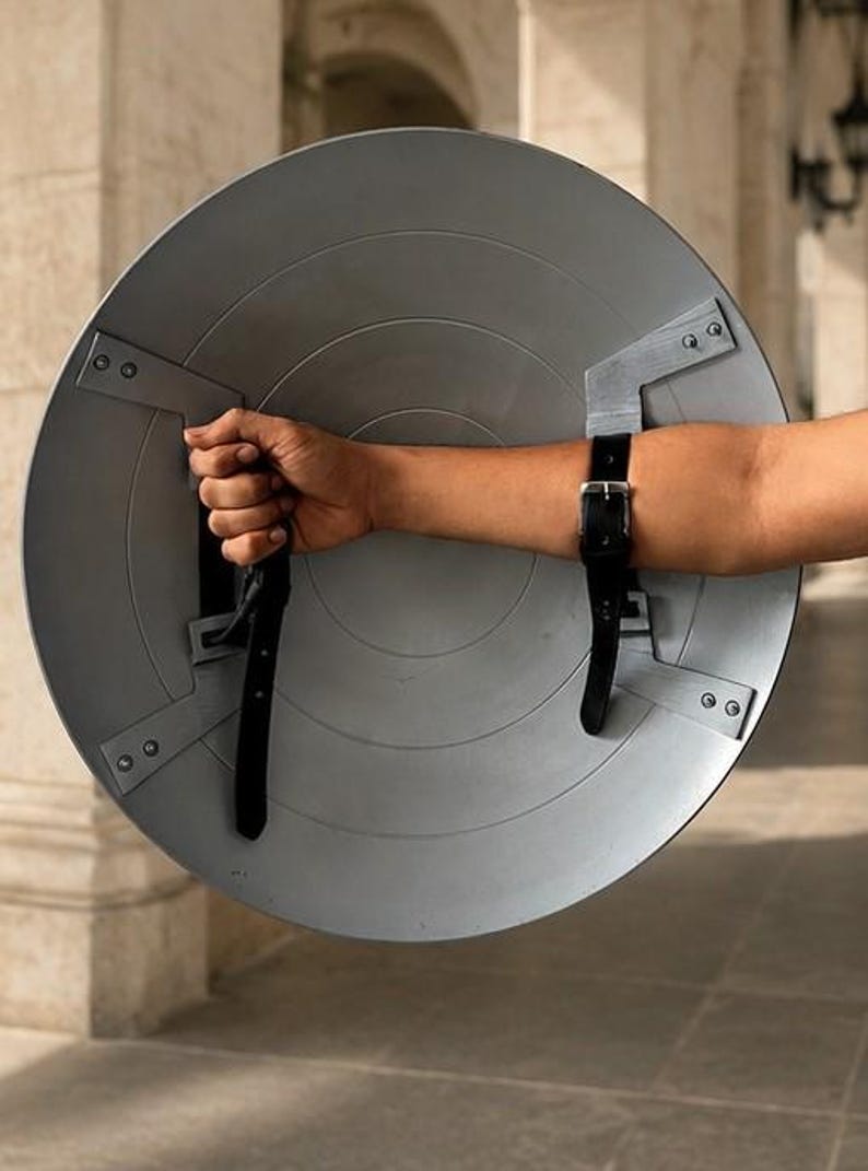 Op de afbeelding: Een grijze, cirkelvormige schild met radiale groeven en metalen beugels. Zwarte leren riemen bevestigen het schild aan de arm. Het schild wordt door een hand vastgehouden, wat duidt op een rekwisiet of kostuumonderdeel.