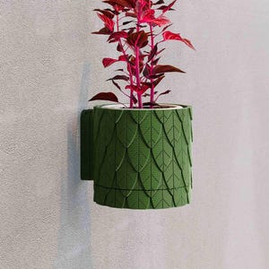 May include: A green wall-mounted planter with a leaf-like pattern, holding a plant with vibrant red leaves. The planter is cylindrical and has a built-in bracket for easy wall mounting. The plant adds a pop of color against the neutral wall.