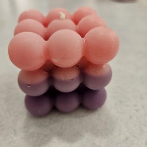 May include: A square, bubble-shaped candle with a gradient of colors. The top layer is a light pink, transitioning to a medium pink, and finally a deep purple at the base. A white wick is visible.