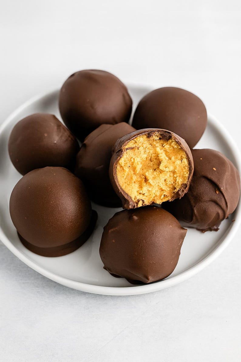 May include: A white plate displays several chocolate-covered peanut butter balls, with one cut open to show the peanut butter filling. The dark brown chocolate coating contrasts with the light yellow-orange peanut butter centre, a delicious treat.