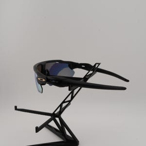 May include: Black sunglasses with blue mirrored lenses are displayed on a black stand. The sunglasses have a sleek, modern design with a curved frame and arms. The lenses reflect a blue hue, and the frame has a small gold accent.