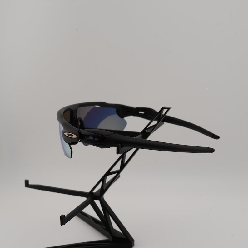 May include: Black sunglasses with blue mirrored lenses are displayed on a black stand. The sunglasses have a sleek, modern design with a curved frame and arms. The lenses reflect a blue hue, and the frame has a small gold accent.