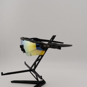 May include: Black sunglasses with a yellow and blue reflective lens. The sunglasses are displayed on a black stand. The lenses have the word "PRIZM" printed on them.