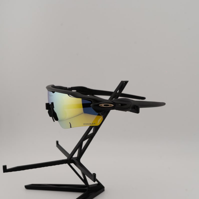 May include: Black sunglasses with a yellow and blue reflective lens. The sunglasses are displayed on a black stand. The lenses have the word "PRIZM" printed on them.