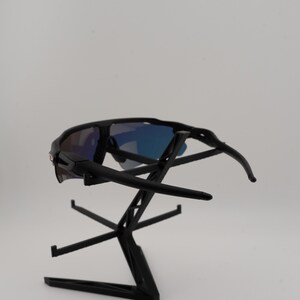 May include: Black sunglasses with blue mirrored lenses are displayed on a black stand. The sunglasses have a sporty design with curved temples and a sleek, modern aesthetic. The lenses reflect a gradient of blue hues.