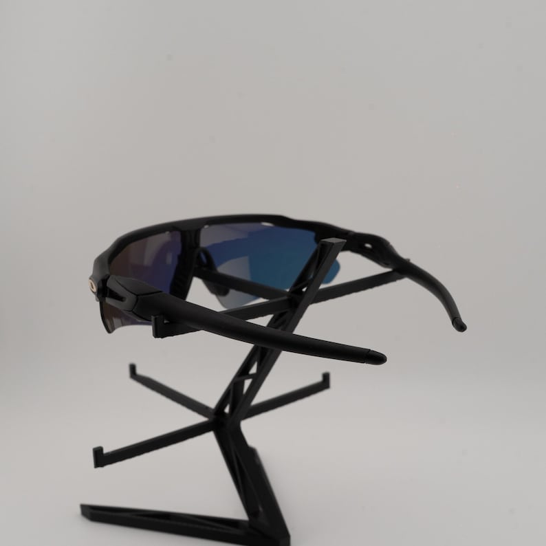 May include: Black sunglasses with blue mirrored lenses are displayed on a black stand. The sunglasses have a sporty design with curved temples and a sleek, modern aesthetic. The lenses reflect a gradient of blue hues.