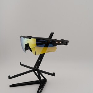 May include: Black Oakley sunglasses with yellow and blue lenses are displayed on a black stand. The sunglasses have a sleek, modern design with the Oakley logo visible on the frame. The lenses have a reflective, mirrored finish.