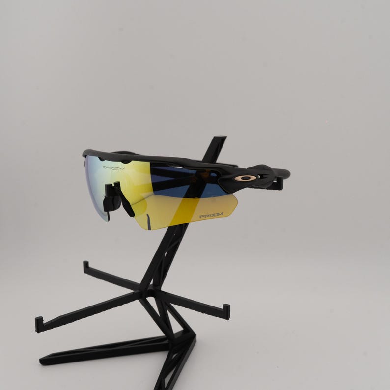 May include: Black Oakley sunglasses with yellow and blue lenses are displayed on a black stand. The sunglasses have a sleek, modern design with the Oakley logo visible on the frame. The lenses have a reflective, mirrored finish.