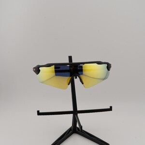 May include: Black sports sunglasses with yellow-tinted lenses. The sunglasses have a sleek, modern design with a black frame and a curved shape. The lenses appear to have a mirrored finish. The sunglasses are displayed on a black stand.