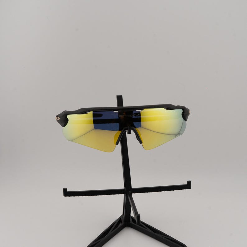 May include: Black sports sunglasses with yellow-tinted lenses. The sunglasses have a sleek, modern design with a black frame and a curved shape. The lenses appear to have a mirrored finish. The sunglasses are displayed on a black stand.