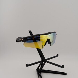 May include: Black sports sunglasses with interchangeable yellow and blue lenses. The sunglasses are displayed on a black stand against a white background. The lenses have a reflective, mirrored finish.