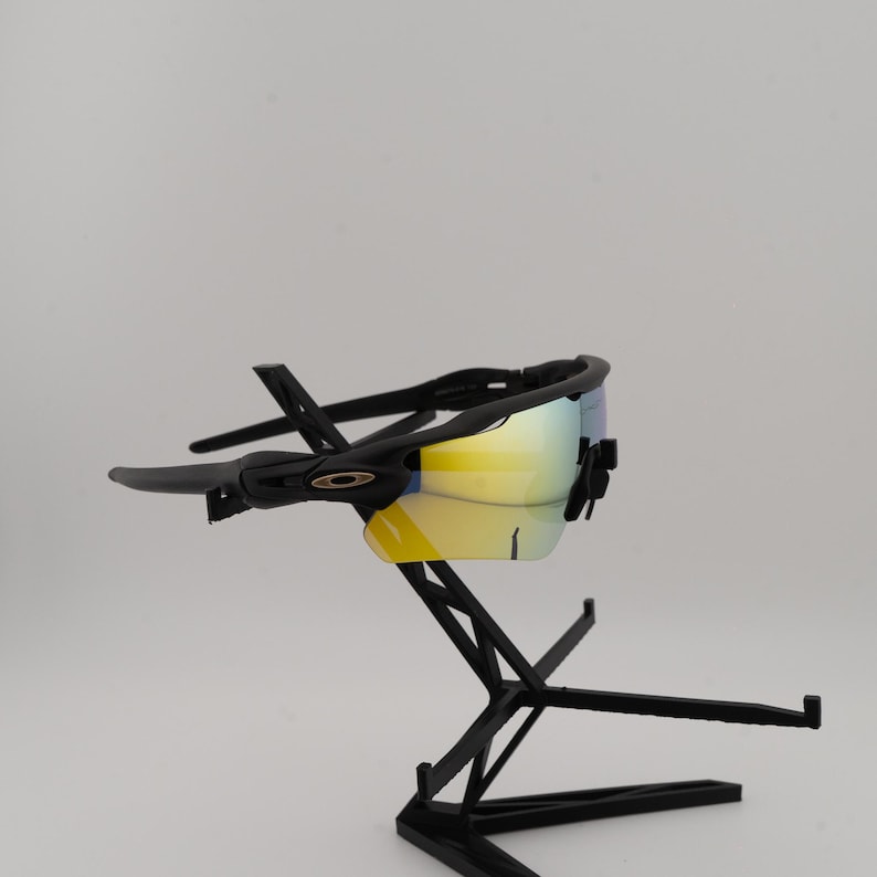 May include: Black sports sunglasses with a yellow and blue mirrored lens. The sunglasses are displayed on a black stand. The frame has a sleek, modern design, suitable for outdoor activities.