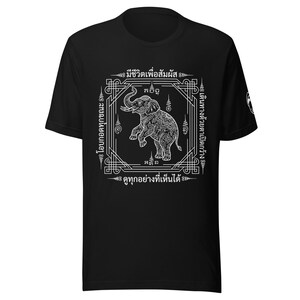 May include: Black t-shirt featuring a white elephant design with intricate patterns and text in a square frame. The design includes Thai script and decorative elements. The shirt has short sleeves.