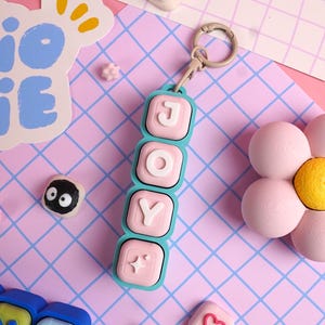 May include: A vertical keychain featuring four square, light blue-framed blocks with rounded corners. The blocks contain white raised letters spelling JOY and a white star symbol on a soft pink background, attached to a beige lobster clasp.