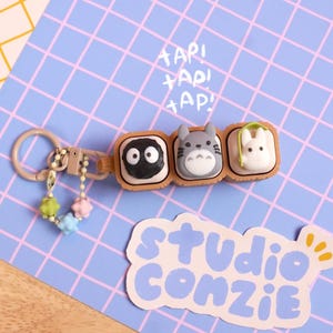 May include: A keychain featuring three square, tan-colored compartments containing small, sculpted figures of a black soot sprite, a grey Totoro, and a white rabbit-like creature. A small chain with colorful star-shaped beads hangs from the ring.