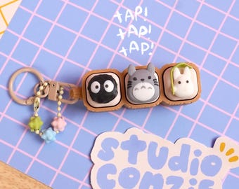 May include: A keychain featuring three square, tan-colored compartments containing small, sculpted figures of a black soot sprite, a grey Totoro, and a white rabbit-like creature. A small chain with colorful star-shaped beads hangs from the ring.