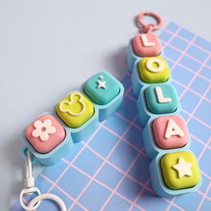 Può includere: Due portachiavi personalizzati composti da blocchi quadrati azzurri con inserti colorati. Uno compone la scritta LOLA con lettere e una stella, mentre l'altro presenta un fiore, il contorno di una testa di topo e un'icona a forma di scintilla.