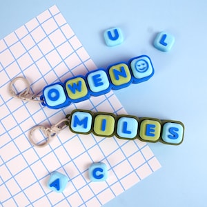 May include: Two silicone keychains featuring square beads with letters spelling OWEN and MILES. The OWEN keychain has blue and green beads with a smiley face, while the MILES keychain has green and light blue beads. Both have metal key rings.