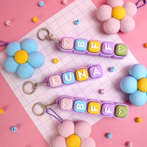 May include: Three keychains featuring pastel-colored square beads with letters and heart symbols. Two keychains spell BFF and one spells LUNA. Each keychain is attached to a large, multi-petaled flower charm in shades of blue or pink.