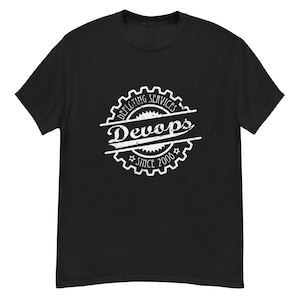 Puede incluir: Camiseta negra con un gráfico blanco. El gráfico presenta la palabra "DevOps" en una fuente estilizada, rodeada por un diseño de engranaje y el texto "Deploying Services Since 2006".