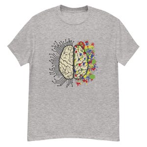 Puede incluir: Camiseta gris con un gráfico de cerebro dividido en dos mitades. Un lado es un diseño de placa de circuito, el otro es una salpicadura de pintura colorida. El gráfico del cerebro está centrado en la camiseta.