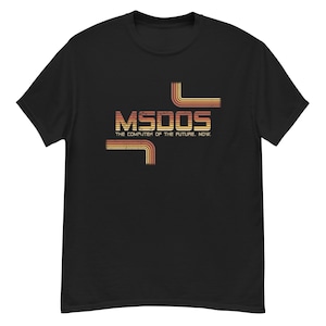 Puede incluir: Camiseta negra con el texto "MSDOS THE COMPUTER OF THE FUTURE. NOW." en una fuente retro. El diseño presenta líneas horizontales y curvas naranjas y amarillas, evocando una estética de ordenador vintage.