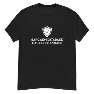 Op de afbeelding: Zwart T-shirt met de witte tekst "SARCASM DATABASE HAS BEEN UPDATED" en een wit schildpictogram met een groene vink. Het T-shirt heeft een ronde hals.