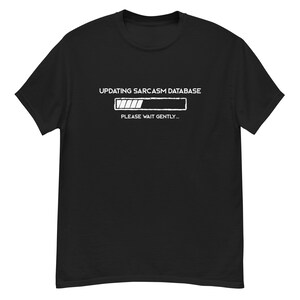 May include: Black t-shirt featuring the text "UPDATING SARCASM DATABASE" above a loading bar graphic, with "PLEASE WAIT GENTLY..." underneath. The design is in white. The t-shirt has a crew neck and short sleeves.