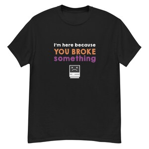 Puede incluir: Camiseta negra con el texto "I'm here because YOU BROKE something" en blanco, naranja y morado. Un gráfico blanco de una computadora triste está debajo del texto. La camiseta es de material suave.