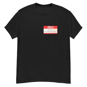 Puede incluir: Camiseta negra con una etiqueta con el texto "HELLO MY NAME IS" en rojo y blanco y "John Doe" con "DROP DATABASE PRODUCTION" debajo. La camiseta es de cuello redondo.