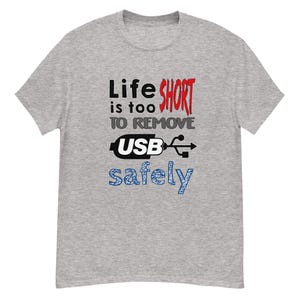 May include: A grey t-shirt featuring the phrase "Life is too short to remove USB safely." The text is in black, red, white, and blue. The shirt has a crew neck and short sleeves.