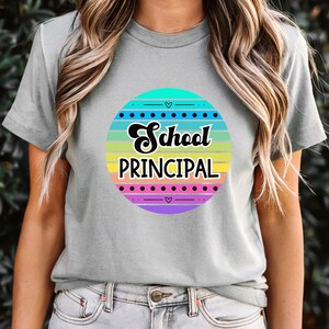 Può includere: T-shirt grigio chiaro con un colorato design circolare. Il design presenta le parole "School PRINCIPAL" in corsivo nero, su strisce orizzontali di vari colori, con piccoli accenti a cuore.