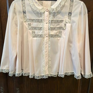 May include: A light pink vintage blouse with long sleeves, a Peter Pan collar, and a button-up front. The blouse features delicate lace trim along the collar, front, and hem. The front has a decorative lace design.