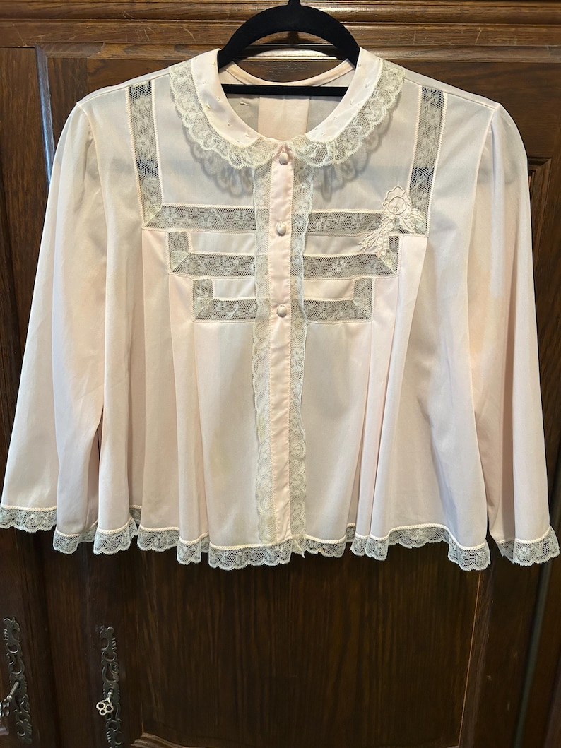 May include: A light pink vintage blouse with long sleeves, a Peter Pan collar, and a button-up front. The blouse features delicate lace trim along the collar, front, and hem. The front has a decorative lace design.